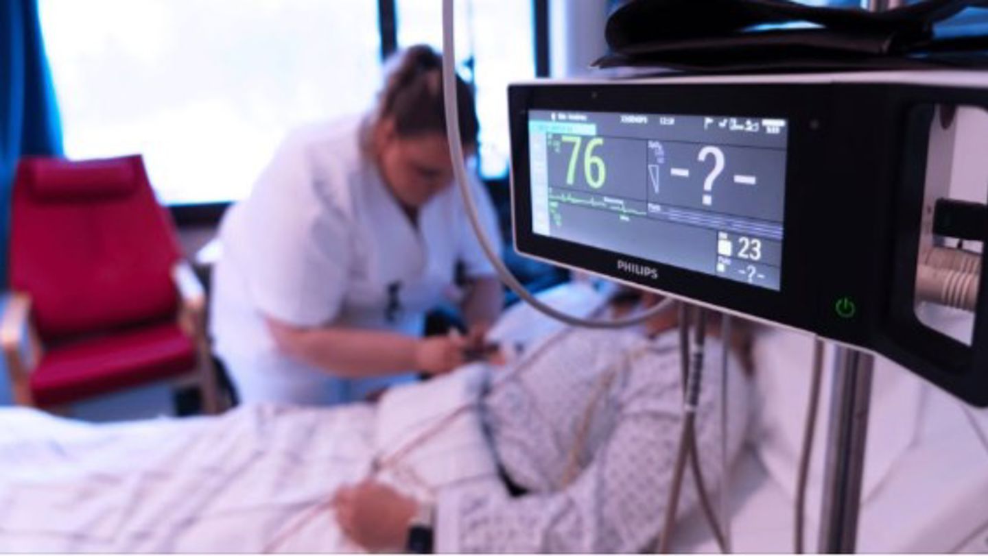 I forgrunnen vises et ekg-apparat på et sykehus og i bakgrunnen sees en pleier bøyd over en pasient i ei seng
