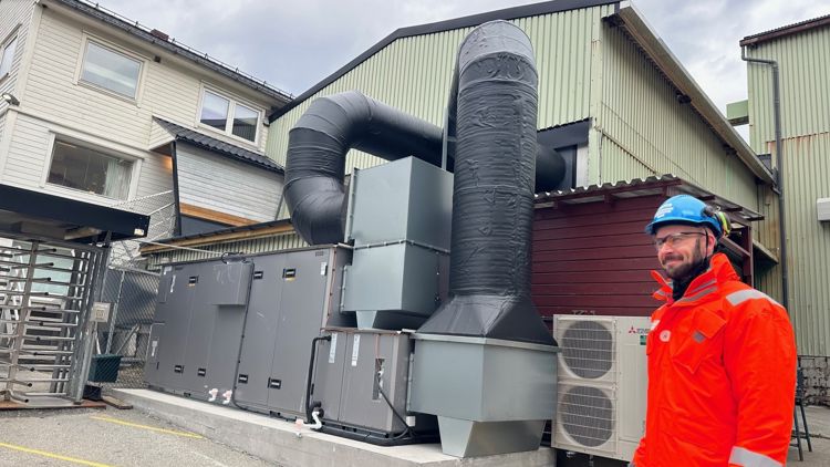 En mann i hjelm og arbeidsjakke står utenfor en industrihall med et stort ventilasjonsanlegg